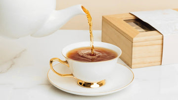 Elegant white teacup with gold trim being filled with tea, set on marble with wooden box nearby