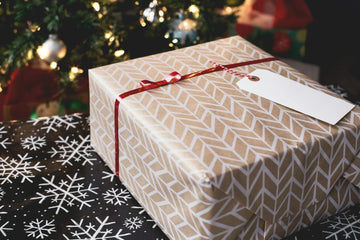 Elegantly wrapped gift with geometric pattern, red ribbon, and blank tag near Christmas tree