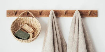 Rustic wooden wall hook with wicker basket holding soap and brush, plus two beige towels, minimalist bathroom decor