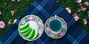 Floral-patterned plate with sliced green apples and teal tea cup on blue plaid blanket amid pink flowers