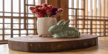 Green ceramic bunny figurine beside vase of dried flowers on rustic wooden tray