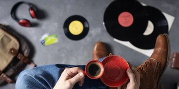 Person holding red coffee cup with vinyl records, headphones, and boots on gray floor — cozy home decor vibe