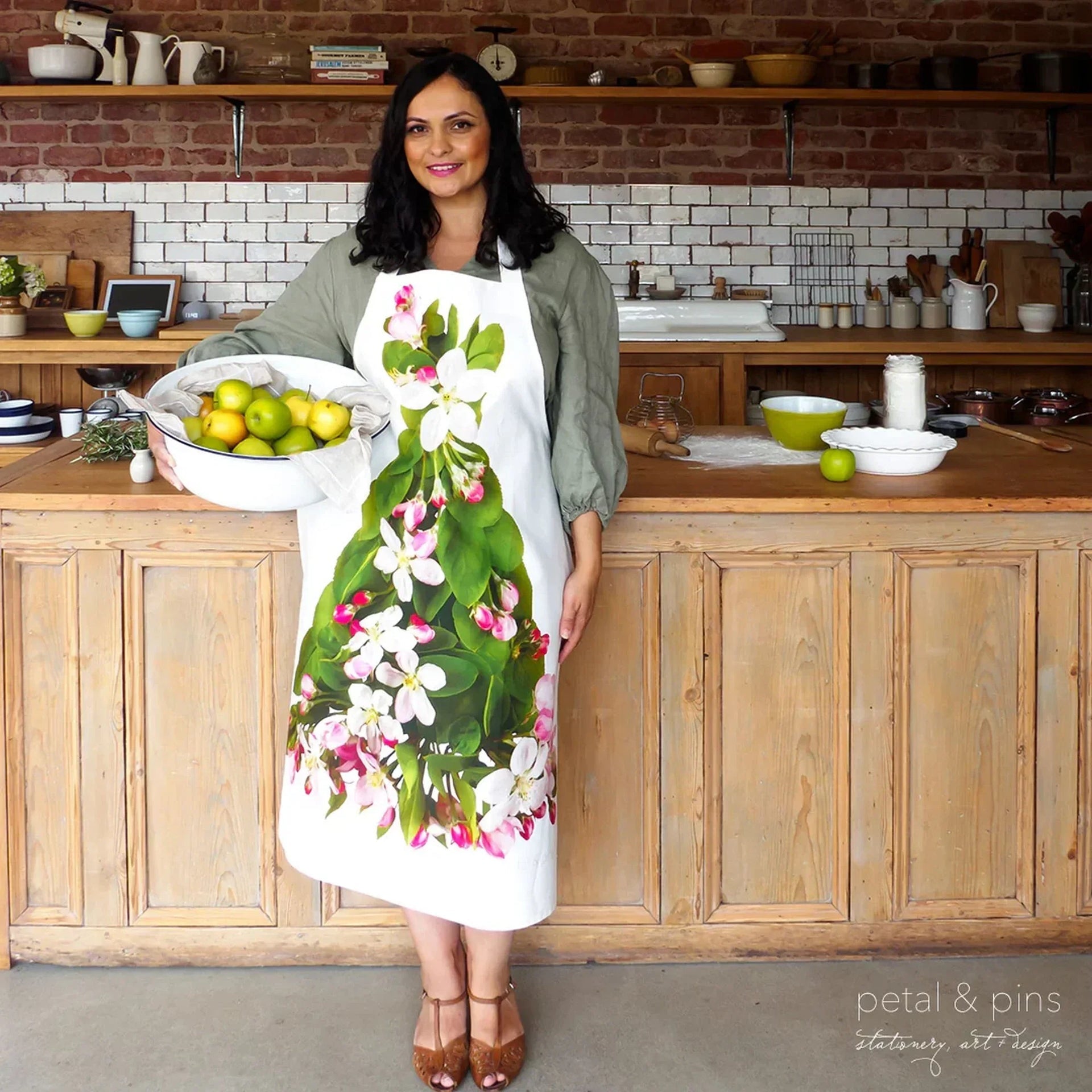 Woman wearing floral apron with apple blossoms, holding fruit bowl in rustic kitchen, home décor accessory