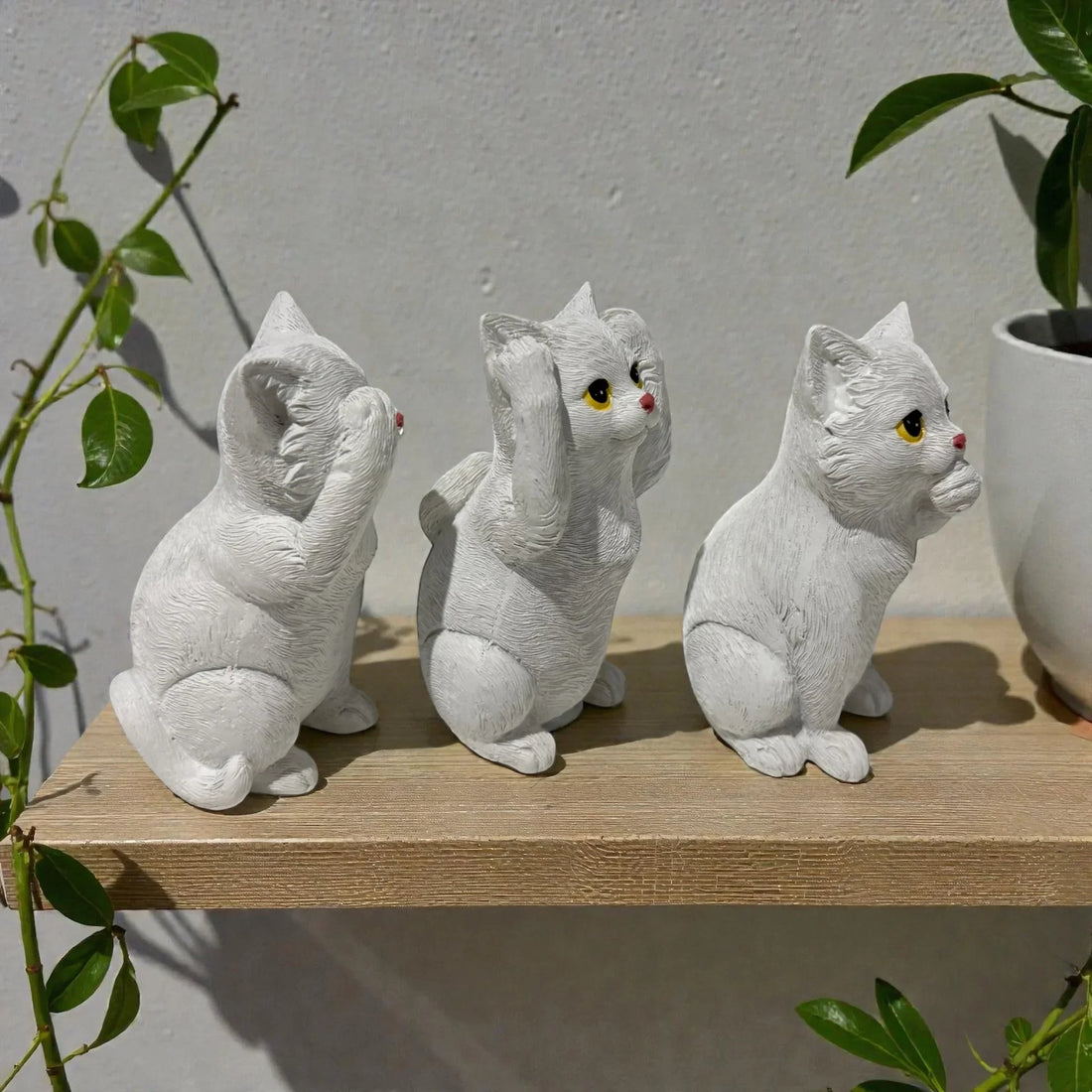 Three white ceramic cat figurines in see-no-evil, hear-no-evil, speak-no-evil poses on wooden shelf