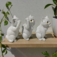 Three white ceramic cat figurines in see-no-evil, hear-no-evil, speak-no-evil poses on wooden shelf