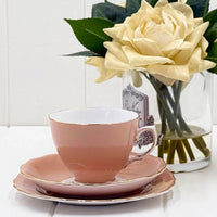 Pink porcelain teacup with gold trim, saucer, and plate set beside cream roses in glass vase, elegant home décor