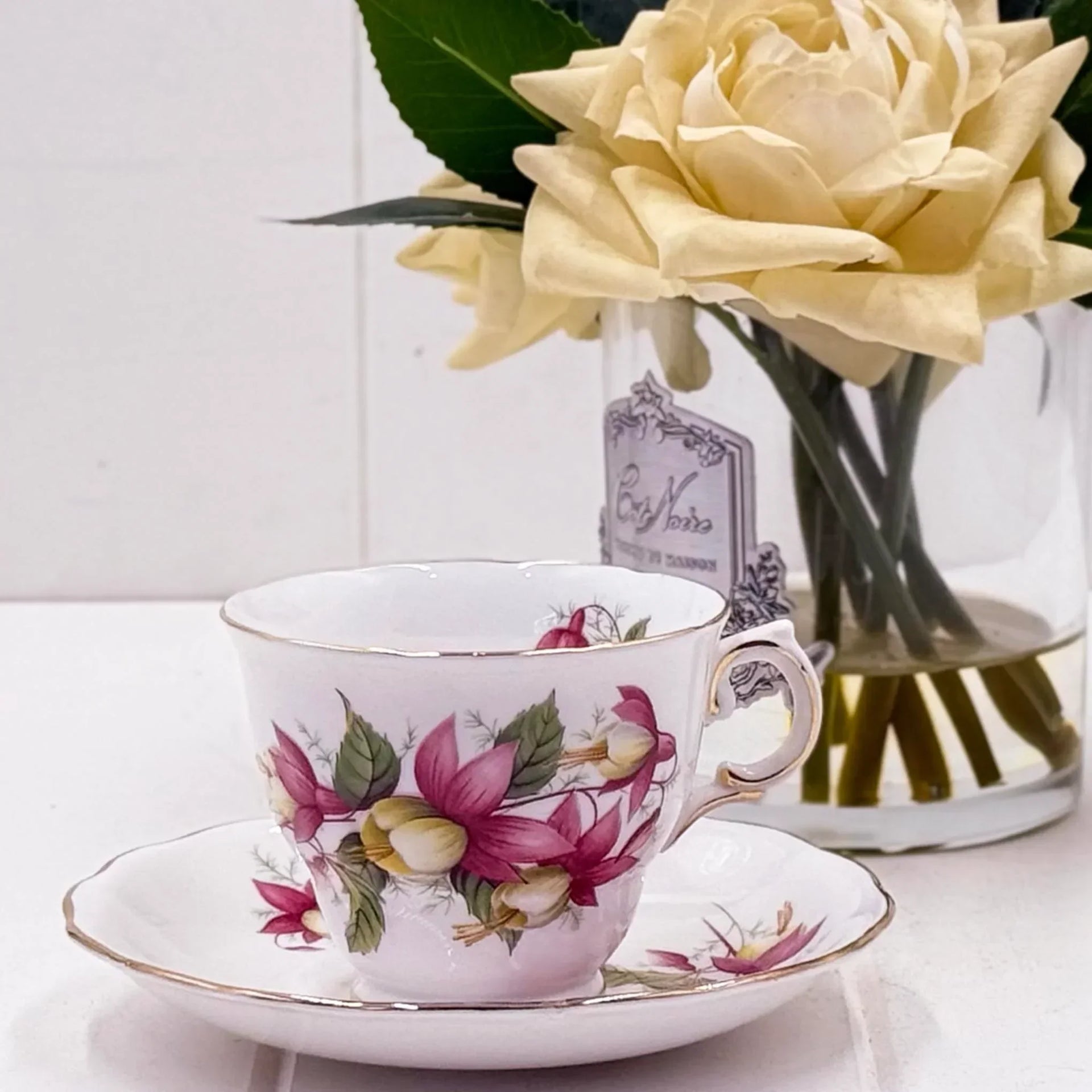 Elegant floral teacup and saucer with gold trim, vintage-style home décor beside a cream rose in vase.