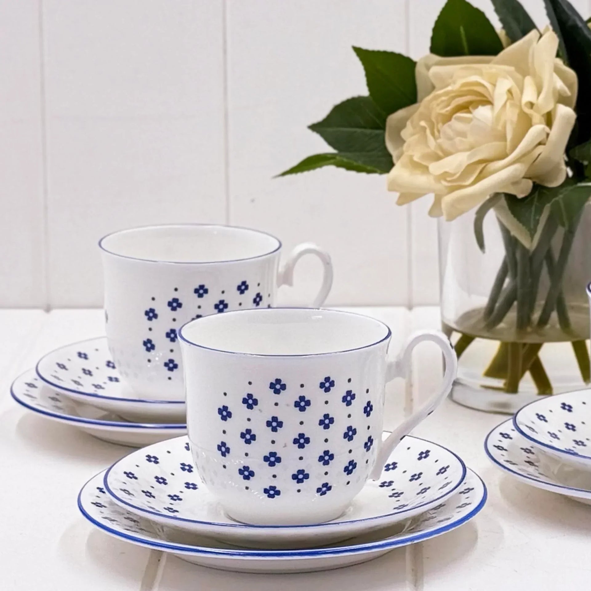 White ceramic teacups with blue floral dots, matching saucers, and a vase of cream roses on a bright table.
