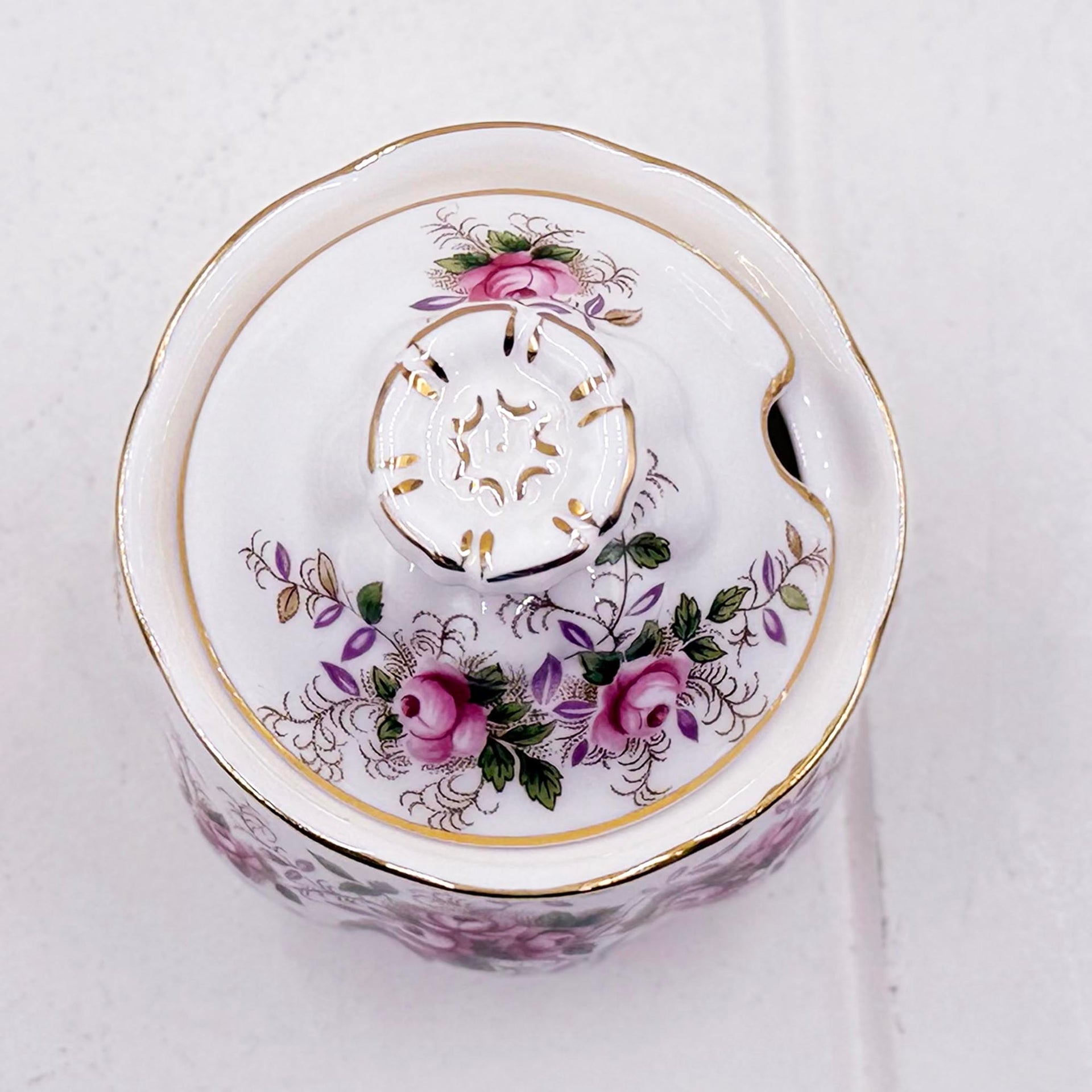 A white Royal Albert  Lavender Rose preserve/sugar/jam pot with a floral design featuring pink roses, green leaves, and gold accents, photographed from a top-down angle on a light textured background.