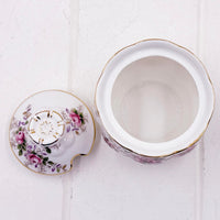 A white Royal Albert lavender Rose bone china sugar pot container with a floral design in pink, purple, and green, accented with gold trim, shown open with its lid placed beside it on a white textured background.