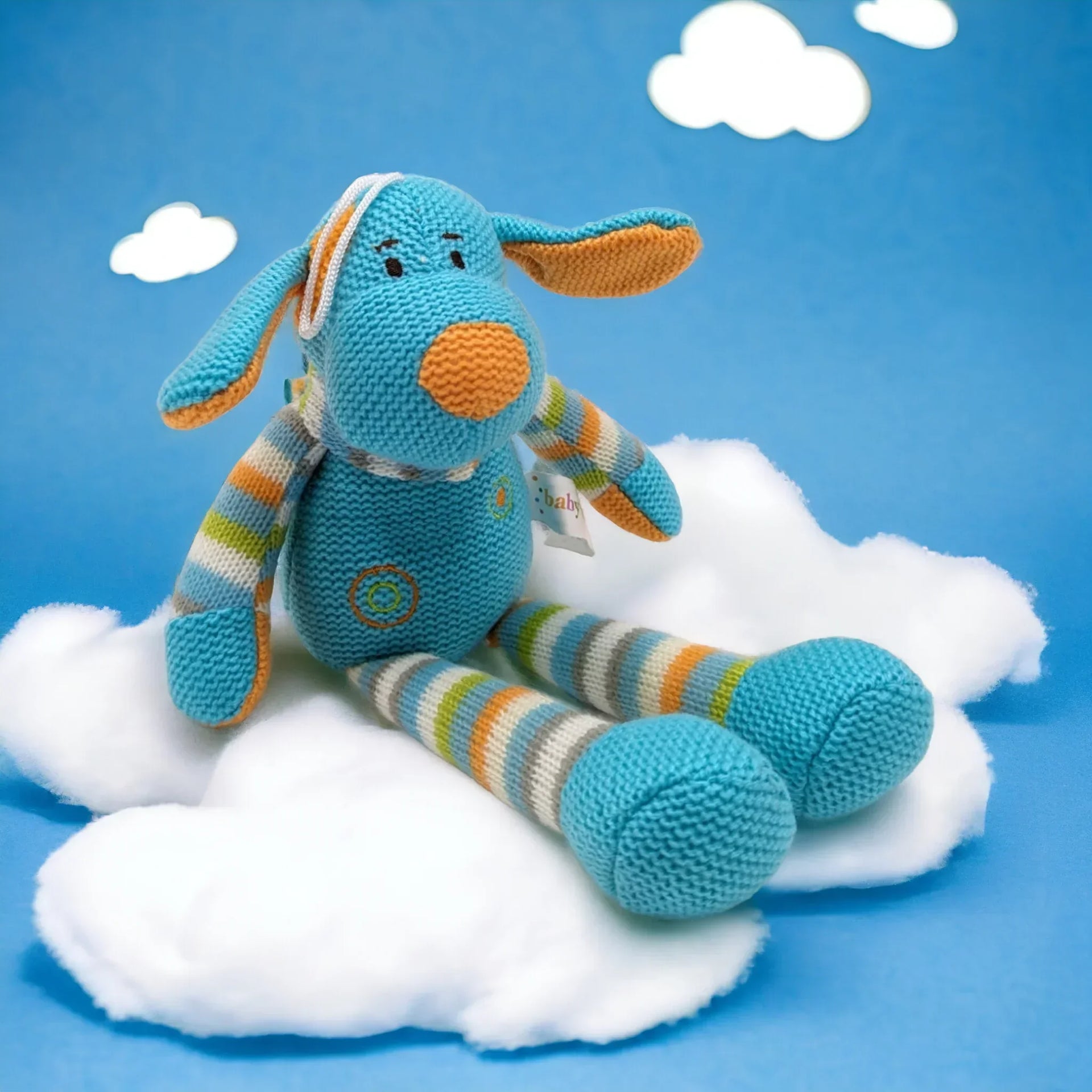 Knitted blue bunny plush toy with striped limbs, sitting on white cloud against blue sky background