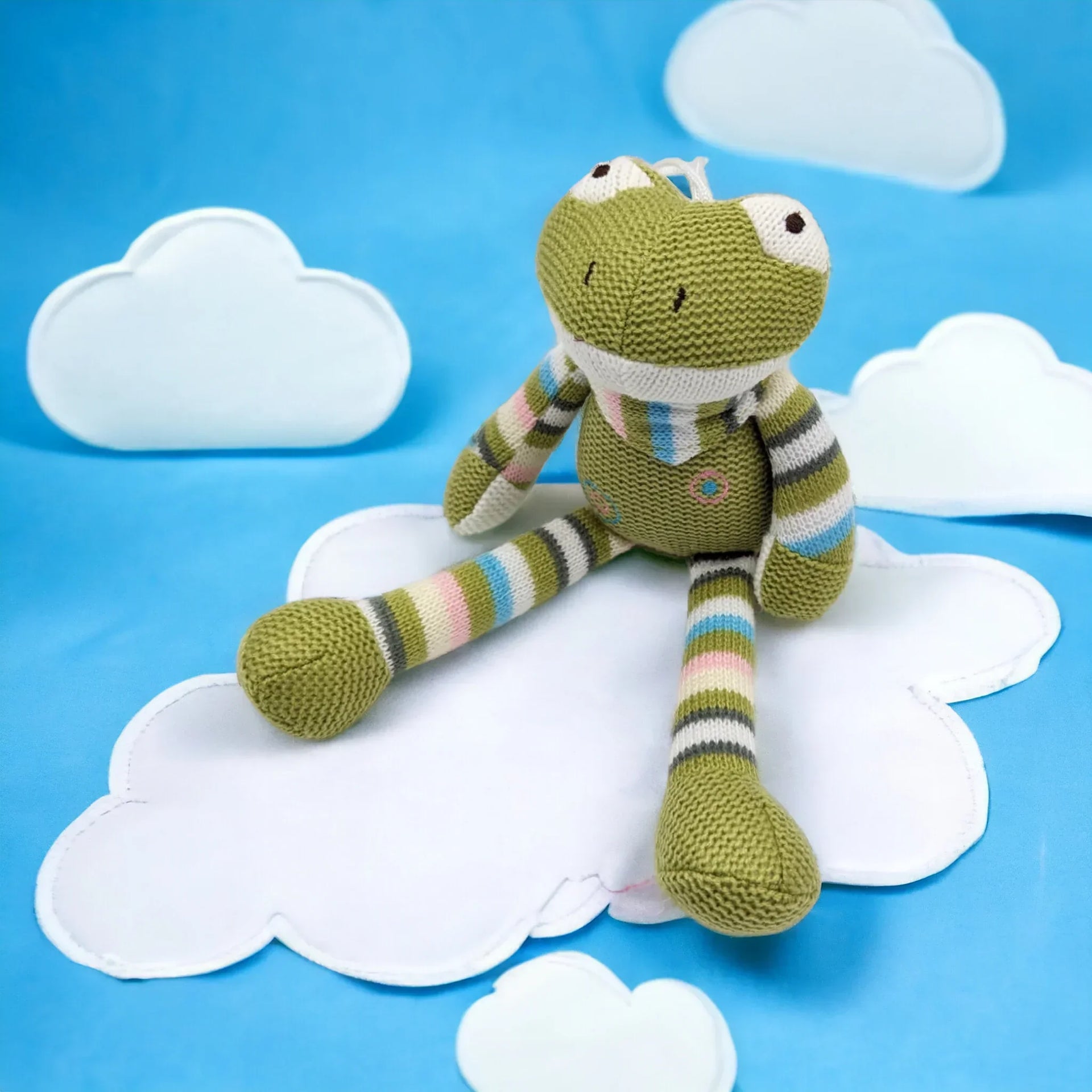Knitted green frog plush toy with striped limbs, sitting on white clouds against blue background