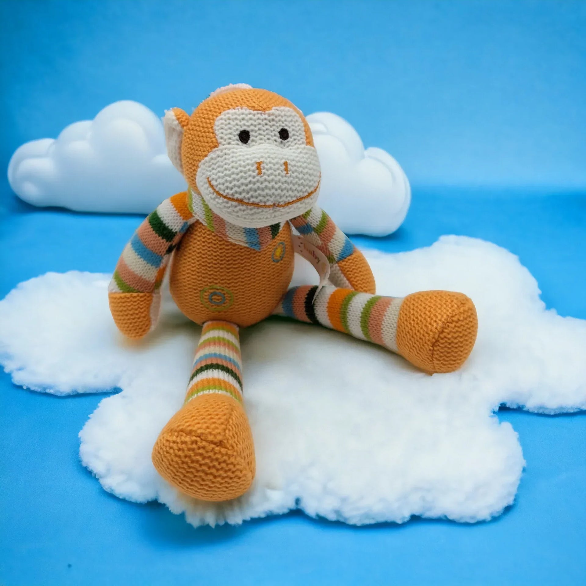 Knitted monkey plush toy with striped limbs, sitting on fluffy white cloud against blue background