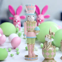 Pastel Easter bunny nutcracker figurine with pink, green, and gold harlequin design, surrounded by decorative eggs and bunnies.