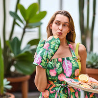 Woman in vibrant vegetable-print apron & oven mitt holding seafood platter, garden backdrop