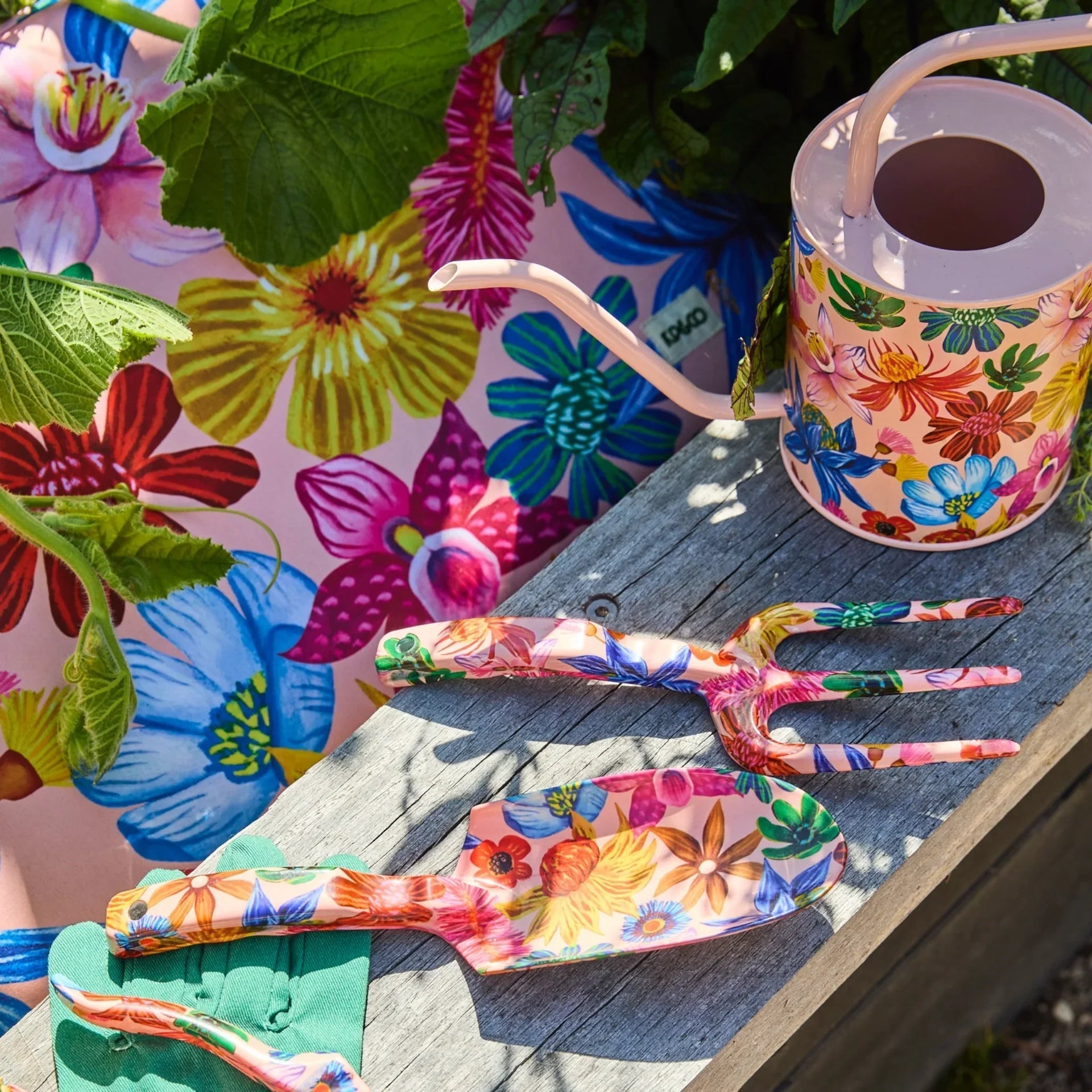Colorful floral garden tools set with watering can, trowel, fork on wooden bench, vibrant blooms backdrop