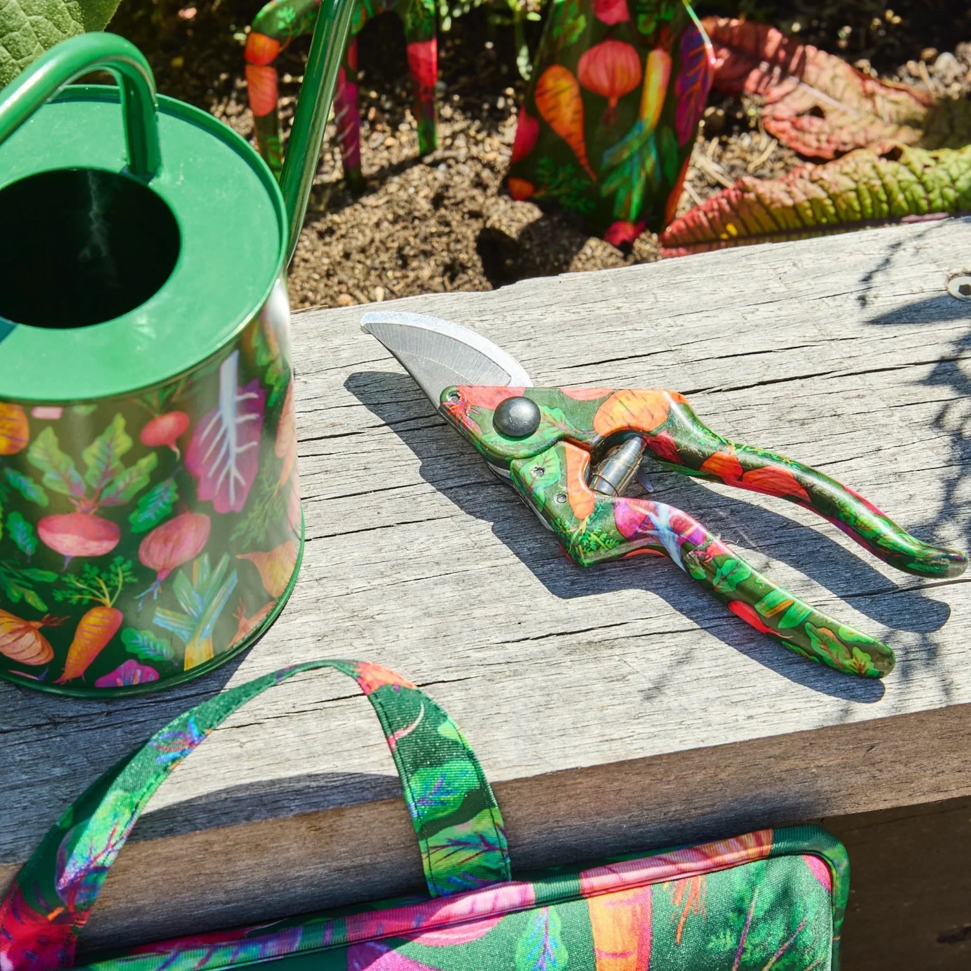 Colorful vegetable-print gardening tools: watering can, pruners, and tote bag on wooden bench