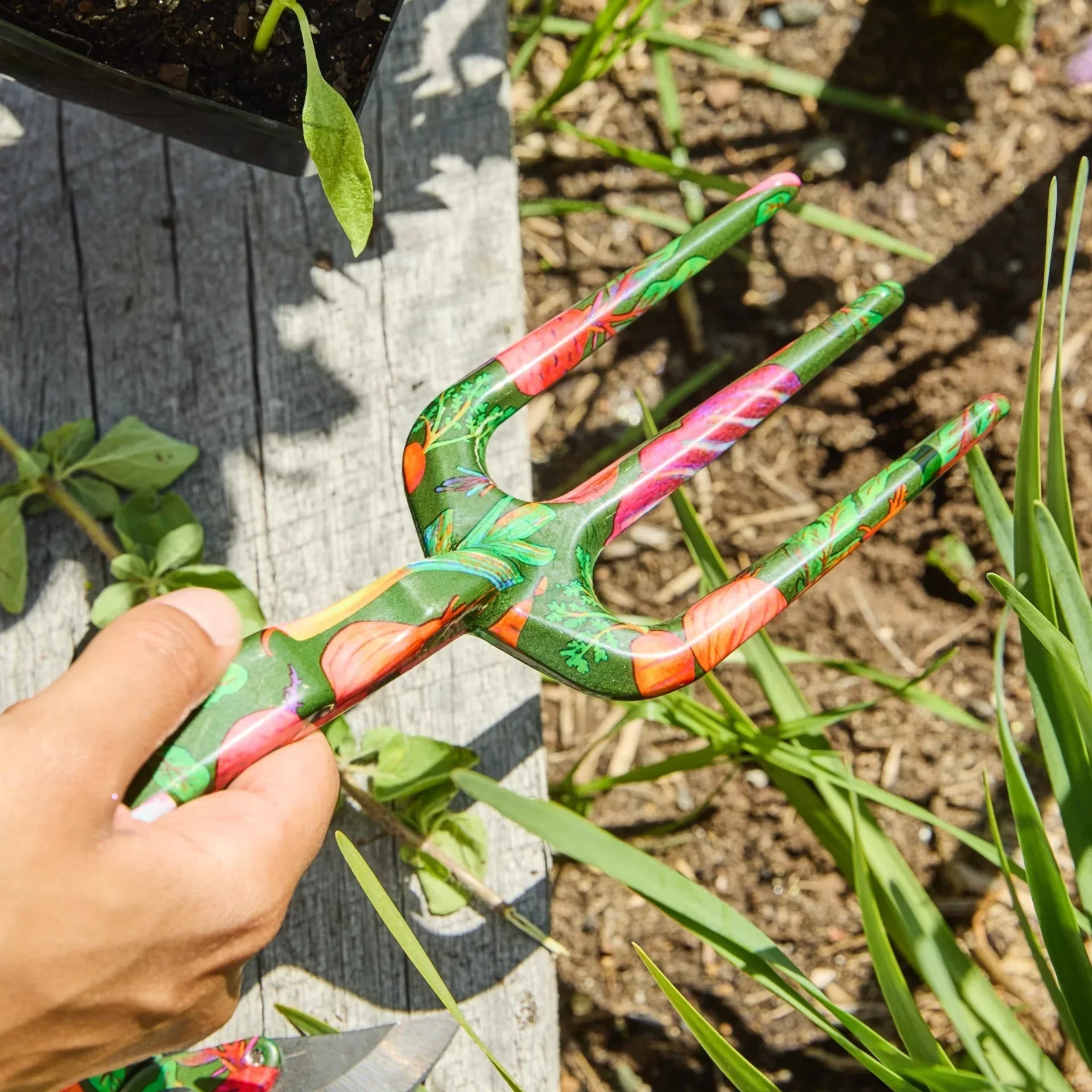 Colorful garden hand fork with floral design, held in garden bed for planting and weeding.