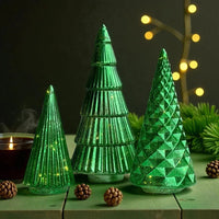 Green glass Christmas tree decorations with LED lights, pinecones, and candle on rustic table