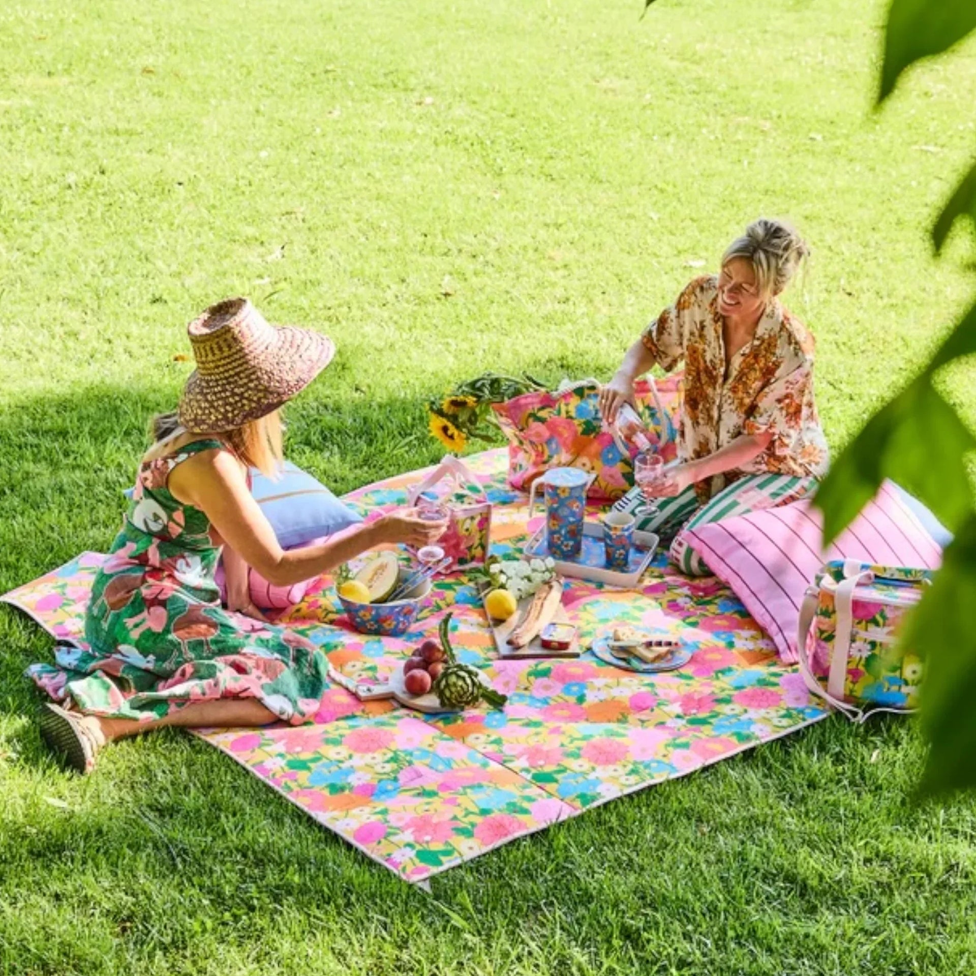 Two women enjoying a picnic on a vibrant floral blanket with matching cooler bags and tableware in a sunny garden.