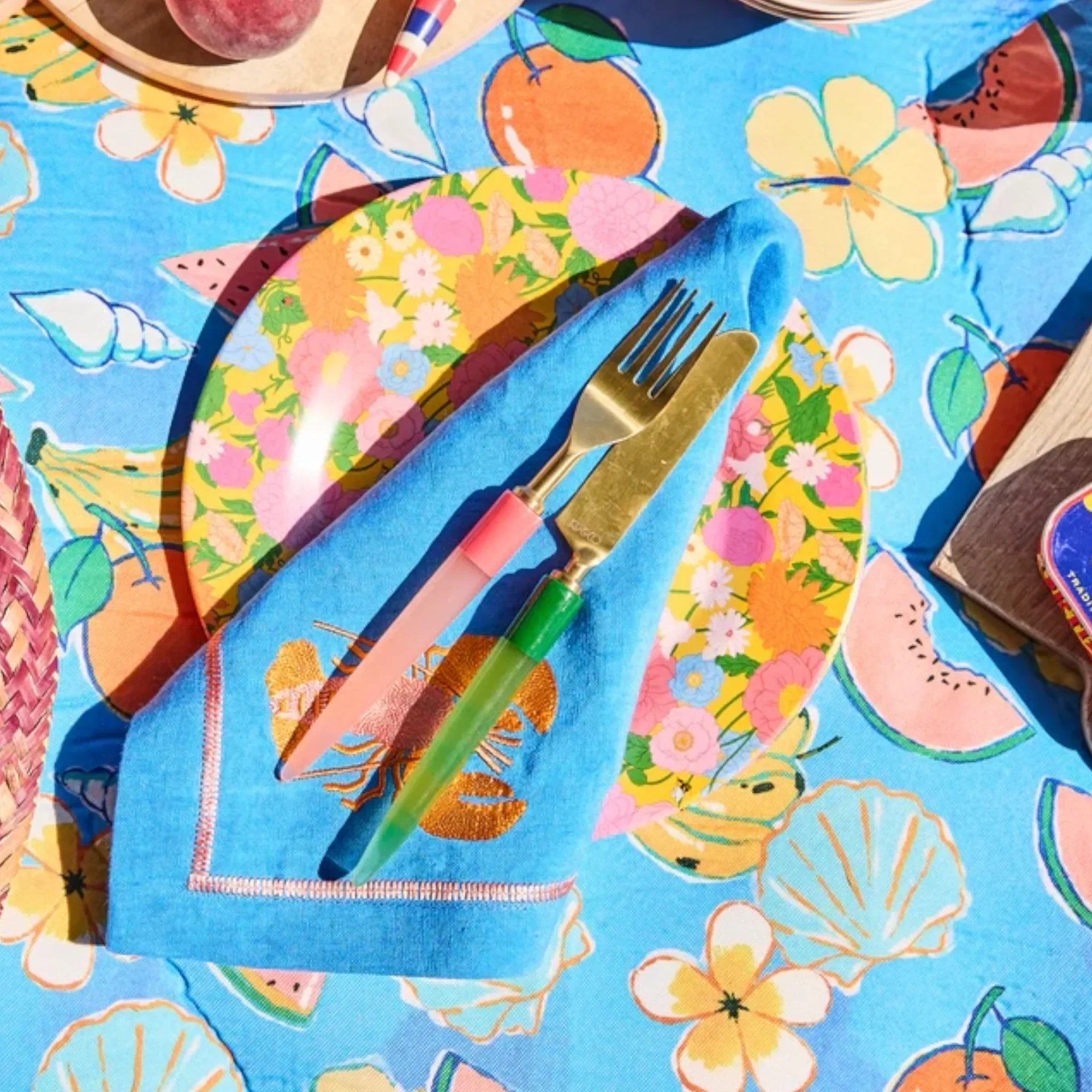 Colorful tropical table setting with floral plates, blue napkin, gold cutlery, and fruit-print tablecloth for summer dining.