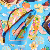 Colorful tropical table setting with floral plates, blue napkin, gold cutlery, and fruit-print tablecloth for summer dining.