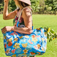 Blue tropical fruit & flower print tote bag with rope handles, carried outdoors by woman in summer hat