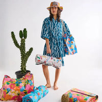 Woman in blue patterned robe holding floral beach bag and rolled towel, with cactus and colorful accessories