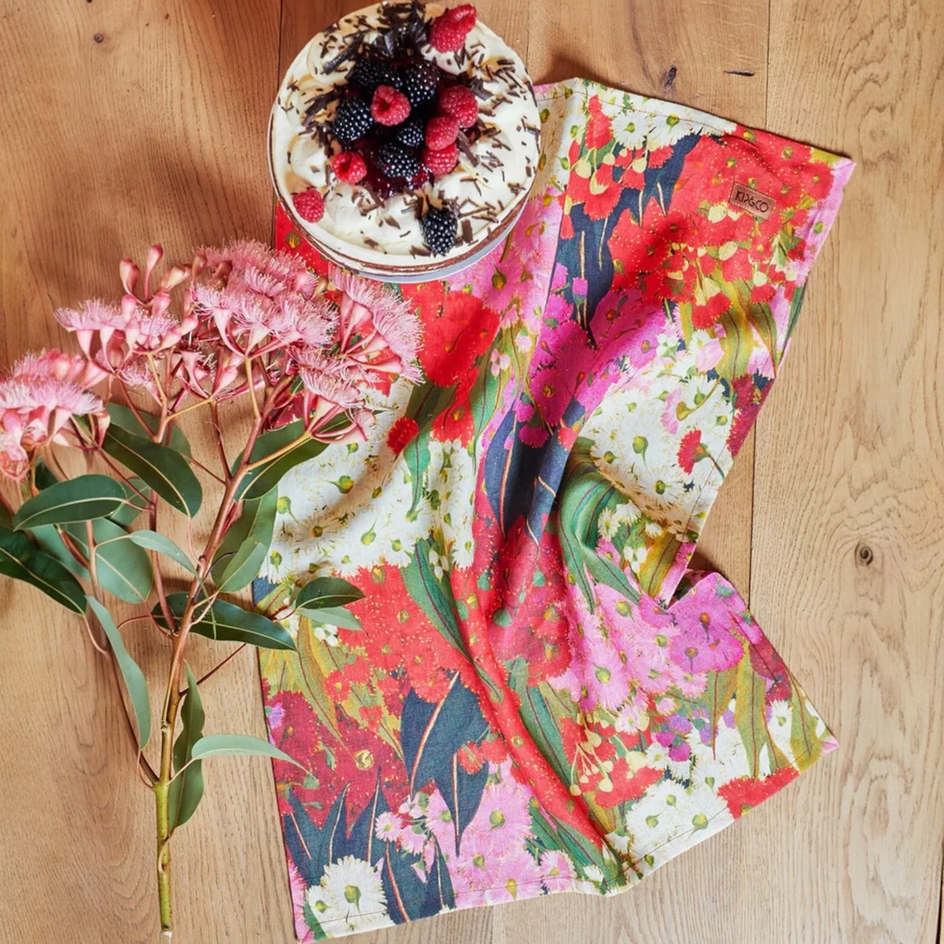 Vibrant floral tea towel with eucalyptus blooms, paired with a berry-topped dessert on wooden table