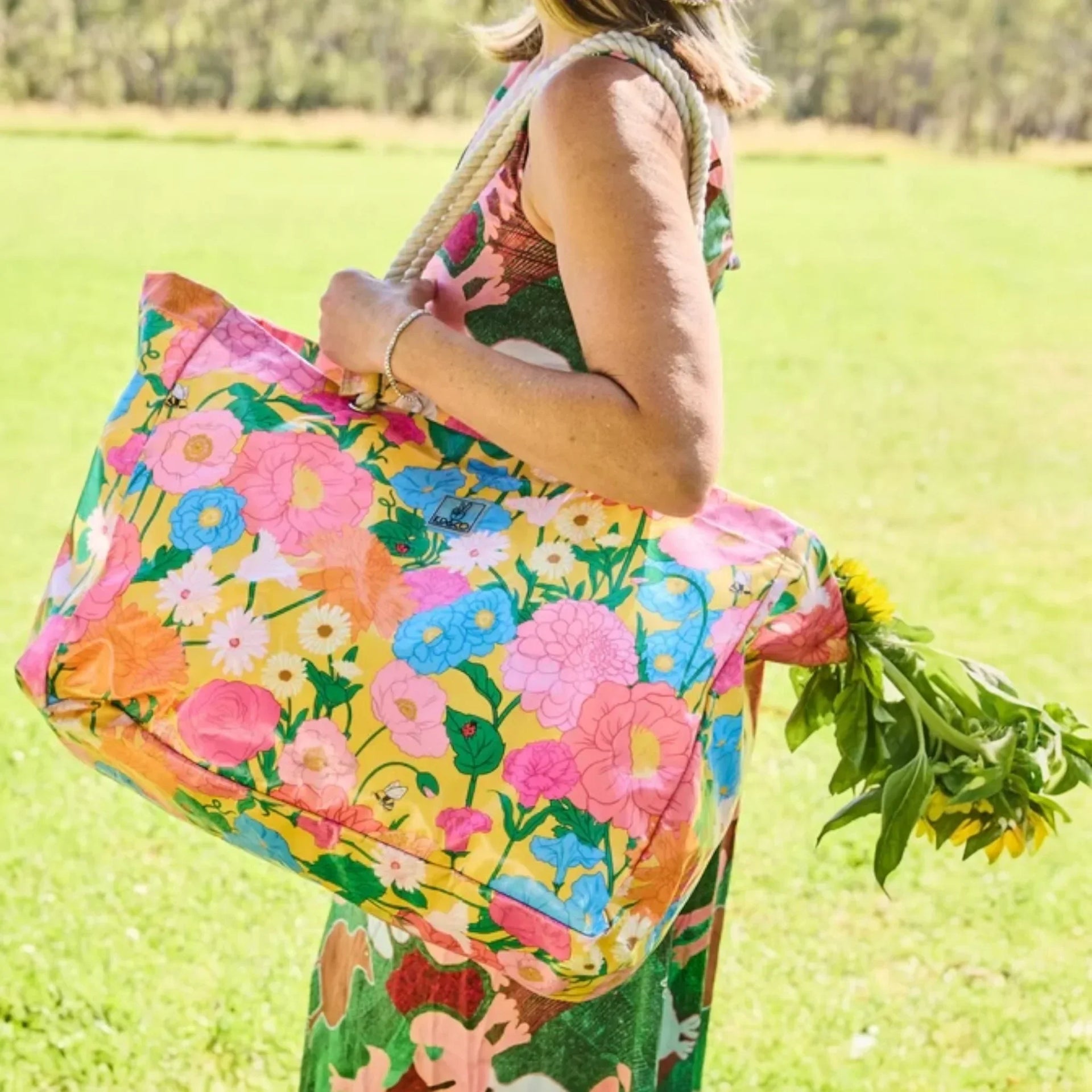 Vibrant floral tote bag with rope handles, carried outdoors with fresh sunflowers, perfect for picnics or market days.