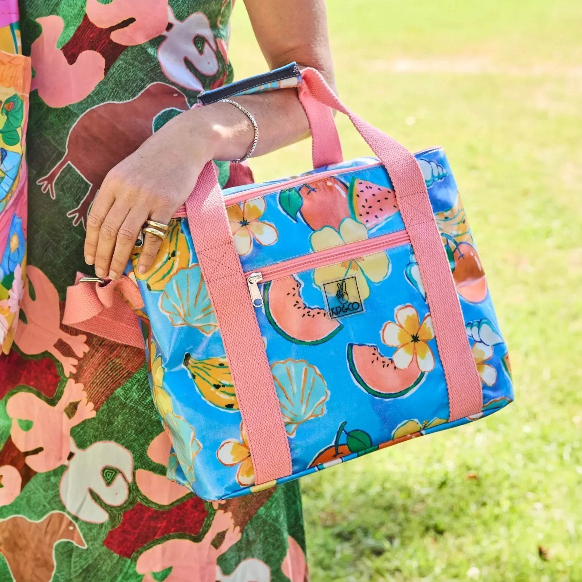 Vibrant blue cooler bag with watermelon, banana & flower print, pink straps, held outdoors