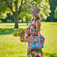 Woman in floral dress & straw hat holding sunflowers, wine, and colorful picnic bags in sunny park