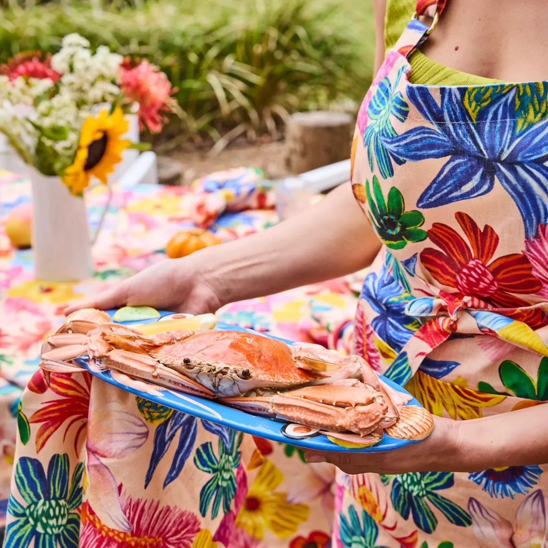 Woman in floral apron holding blue tray with cooked crab, outdoor table setting with sunflowers