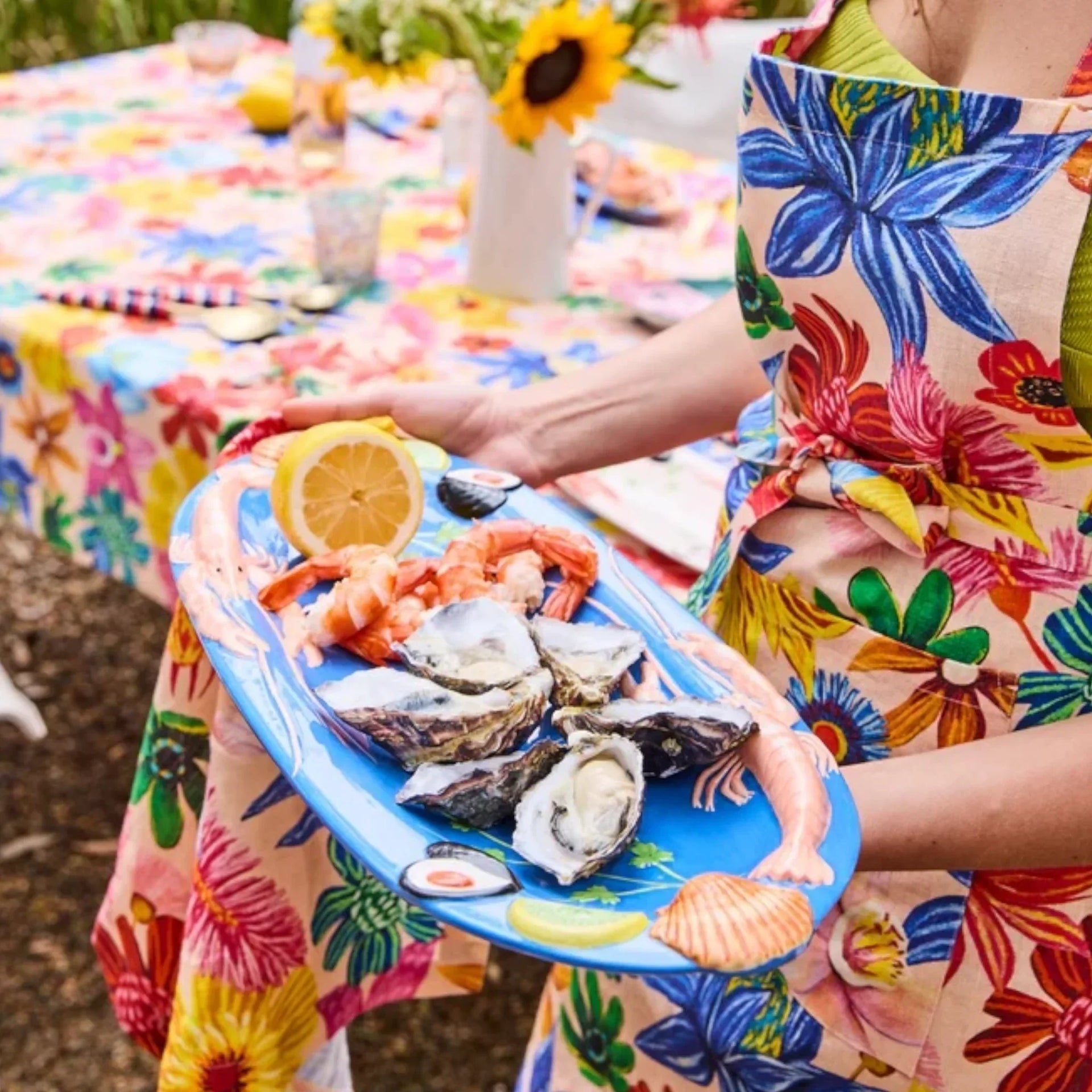 Vibrant floral apron holder serving oysters, prawns, and lemon on a blue seafood platter at a garden table setting.