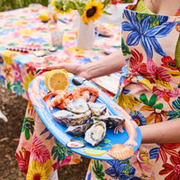 Vibrant floral apron holder serving oysters, prawns, and lemon on a blue seafood platter at a garden table setting.