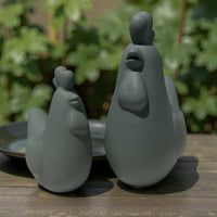 Grey ceramic chicken figurines for home décor, styled on wooden table with greenery background