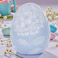 Light blue ceramic Easter egg with embossed hydrangea flowers and leaves, surrounded by pastel eggs and spring blooms.