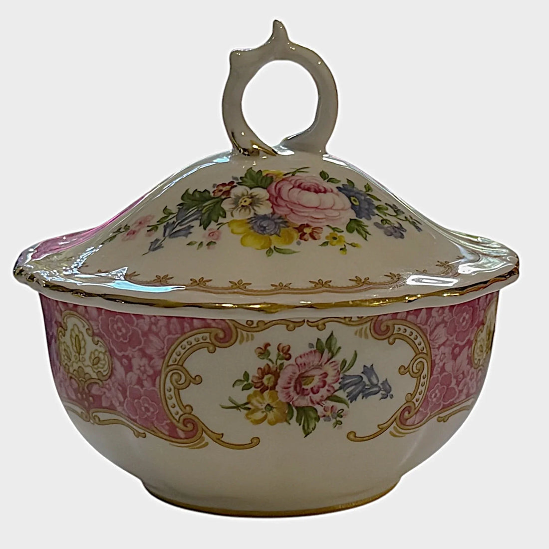 An ornate porcelain tureen with a lid and decorative handle, featuring a pink damask pattern, gold scrollwork, and colorful floral motifs on a cream background, set against a plain light gray backdrop.