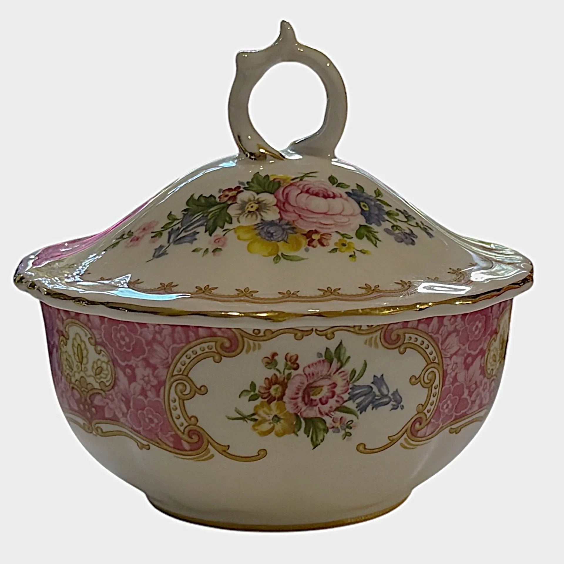 An ornate porcelain tureen with a lid and decorative handle, featuring a pink damask pattern, gold scrollwork, and colorful floral motifs on a cream background, set against a plain light gray backdrop.