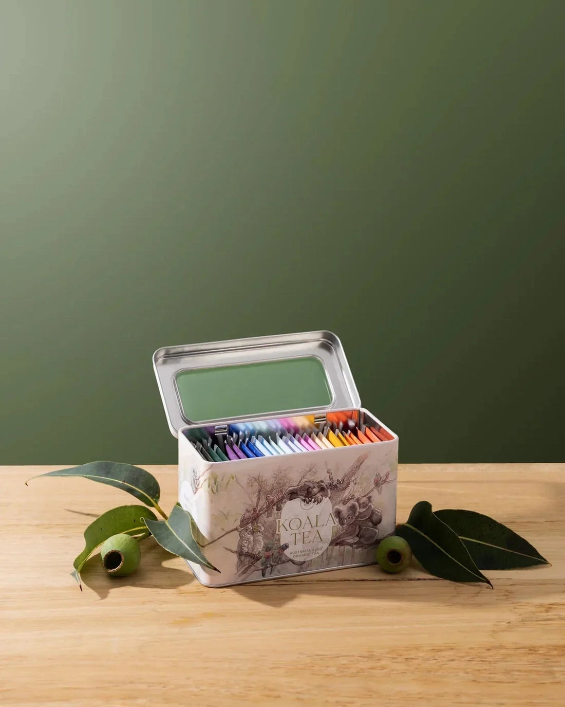 Koala Tea tin with colorful tea bags, eucalyptus leaves on wood, green background
