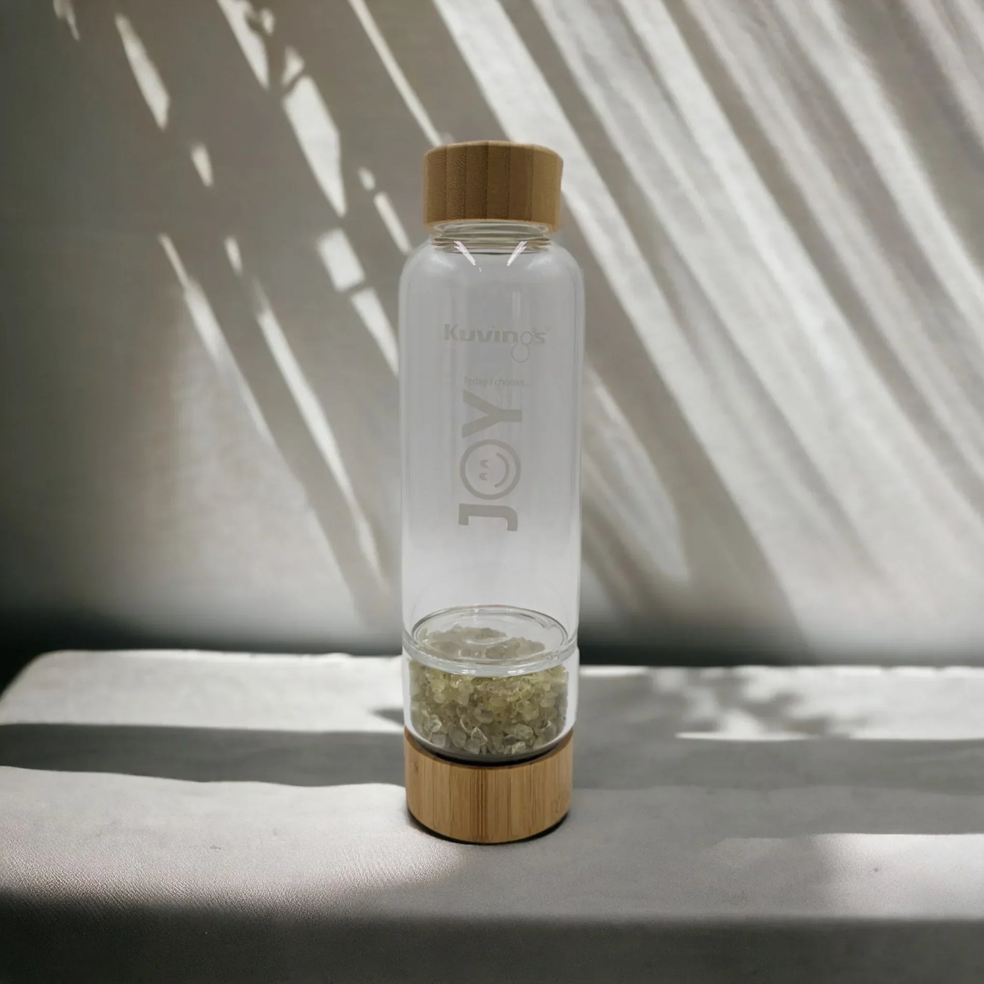 Clear glass water bottle with bamboo lid and base, featuring 'JOY' etching and crystal infuser, styled with leaf shadows.