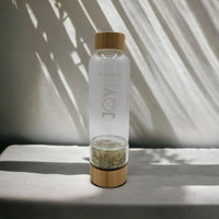 Clear glass water bottle with bamboo lid and base, featuring 'JOY' etching and crystal infuser, styled with leaf shadows.