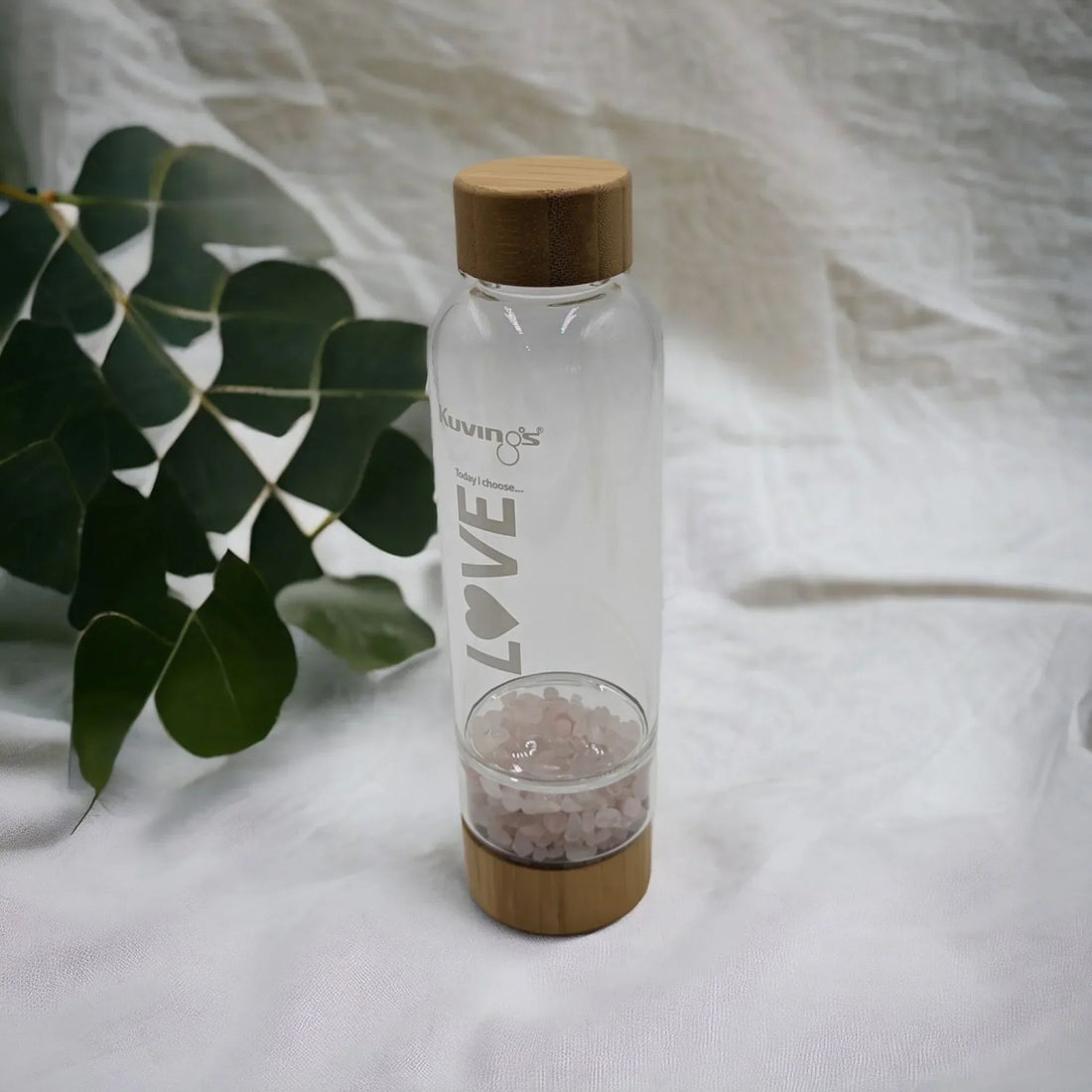 Clear glass water bottle with bamboo lid and pink crystal infuser, 'LOVE' text, beside green leaves on white fabric