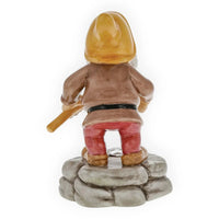 Ceramic garden gnome figurine with yellow hat, red pants, holding tool, standing on stone base